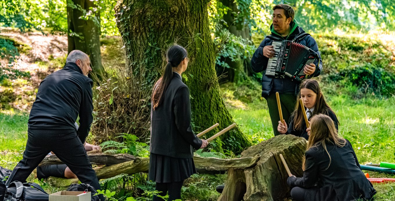 2 adults and 3 students in a sun dappled forest, 1 adult playing the accordion, 3 students playing a tree stump with drum sticks.