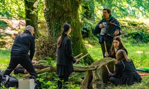 2 adults and 3 students in a sun dappled forest, 1 adult playing the accordion, 3 students playing a tree stump with drum sticks.